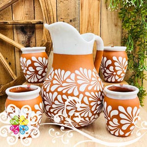 May include: A terracotta pitcher and matching cups set. The pitcher has a white top and handle, with a floral design in white. The cups have a similar design. The set is displayed on a wooden surface.