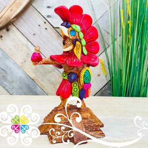 May include: A colorful ceramic figurine of a rooster wearing a red headdress and holding a small figure in its hand. The rooster is standing on a brown pyramid-shaped base with intricate carvings. The figurine is painted in bright colors, including red, yellow, green, blue, and white.