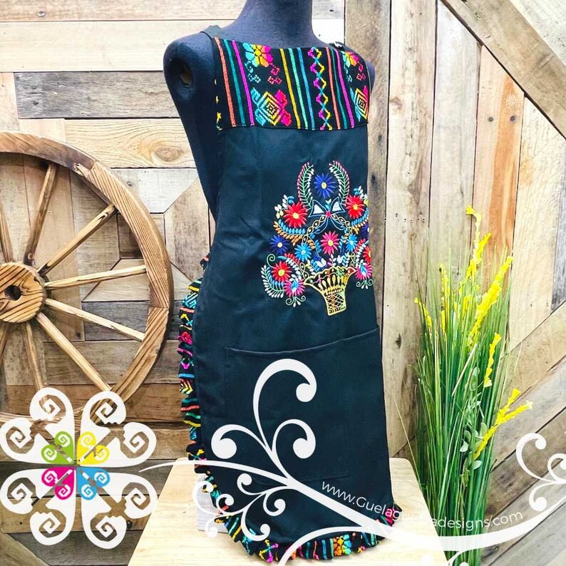 May include: Black apron with a colorful floral embroidery design and a ruffled bottom. The apron has a pocket and a colorful geometric pattern at the top. The text "www.GueladeDesigns.com" is visible at the bottom.