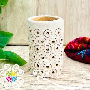 May include: A cylindrical, off-white ceramic vase with a textured surface and a pattern of evenly spaced, circular cutouts. The interior is a warm beige. The vase is displayed on a light wooden surface, with a colorful textile in the background.