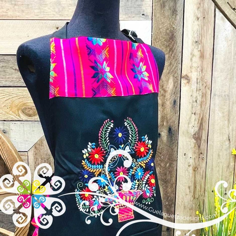 May include: Black apron with a pink and blue embroidered design. The apron has a floral pattern with a basket in the center. The text "www.GuelaguetzaDesigns.com" is embroidered on the bottom right corner.
