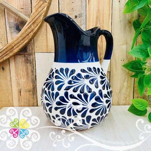 May include: A ceramic pitcher with a navy blue top and handle, transitioning to white with a blue floral design. The pitcher has a curved spout and a rounded body. The design is reminiscent of traditional pottery.