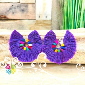 May include: A pair of vibrant purple tassel earrings. Each earring features a fan-shaped design with a colorful beaded center. The earrings are suspended from silver-colored hooks, offering a bold and artistic statement.