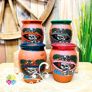 May include: Four terracotta ceramic mugs with colorful Day of the Dead skull designs. Each mug features a skull wearing a hat with flowers and colorful accents. The mugs are perfect for serving beverages.