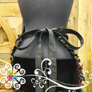 May include: Black apron with a colorful, fringed trim and a black ribbon tie. The apron is tied around a mannequin. The text "www.GuelaguetzaDesigns.com" is visible in the bottom right corner.