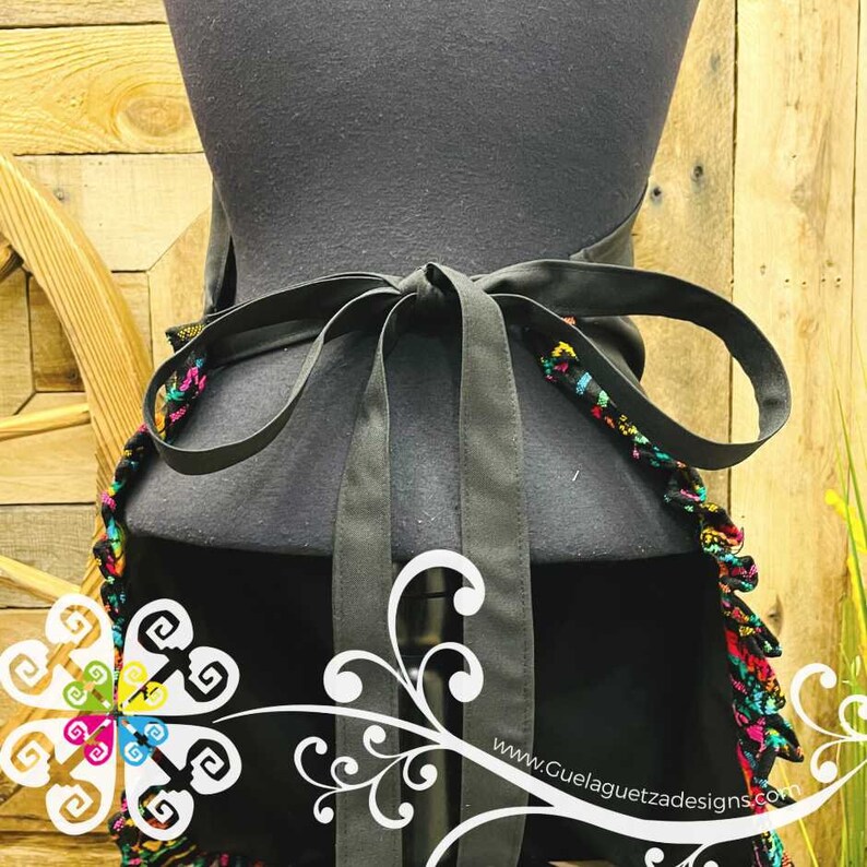 May include: Black apron with a colorful, fringed trim and a black ribbon tie. The apron is tied around a mannequin. The text "www.GuelaguetzaDesigns.com" is visible in the bottom right corner.