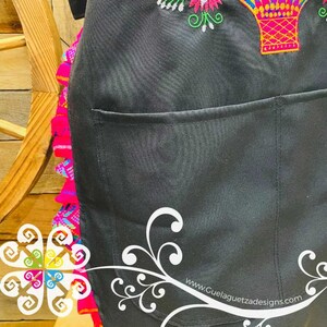 May include: Black apron with a large pocket and colorful embroidery of a hot air balloon and flowers. The apron has a ruffled edge with pink, blue, and green accents. The text "www.GuelaguetzaDesigns.com" is printed on the bottom right corner.