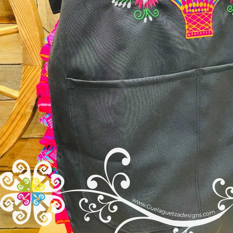 May include: Black apron with a large pocket and colorful embroidery of a hot air balloon and flowers. The apron has a ruffled edge with pink, blue, and green accents. The text "www.GuelaguetzaDesigns.com" is printed on the bottom right corner.