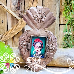 May include: A decorative metal frame in a heart shape, featuring a portrait of Frida Kahlo. The frame has a bronze finish with floral embellishments. The portrait shows Frida with a red flower in her hair.