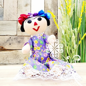 May include: A purple and yellow floral dress with white lace trim, worn by a handmade doll with black hair, red and blue ribbons, and a painted face.
