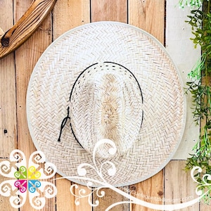 Könnte beinhalten: Ein gewebter Strohhut mit breiter Krempe und einem schwarzen Band um den Kronenbereich. Der Hut wird vor einem Holzgrund präsentiert. Das Design des Hutes ist schlicht und klassisch, geeignet für Sonnenschutz.