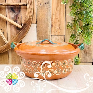 May include: A terracotta cooking pot with a lid, featuring white and green painted designs. The pot has a handle on the lid and side handles. The background includes a wooden surface and decorative elements.