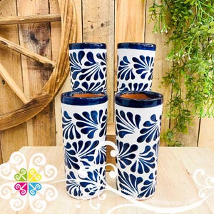 May include: Four white ceramic tumblers with a blue floral design. The rims are dark blue, and the interior is a natural clay color. The tumblers are arranged on a wooden surface, with a wooden wheel and greenery in the background.