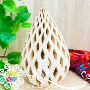 May include: White, teardrop-shaped decorative object with a lattice design. The object has a cream-colored interior visible through the diamond-shaped cutouts. The background includes green leaves and a colorful textile.
