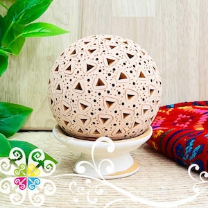 May include: A beige ceramic sphere with triangular cutouts and small dot perforations, resting on a white pedestal with a gold trim. The sphere is a decorative item, possibly a candle holder or diffuser.