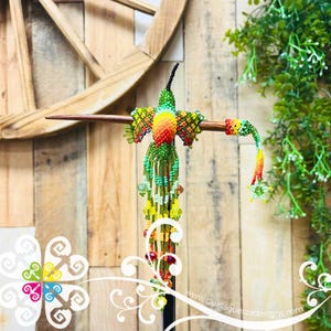 May include: A handcrafted beaded hummingbird sculpture with vibrant colors. The bird features a green body, a yellow and orange chest, and a long, beaded tail. It is mounted on a wooden dowel, with a wooden wheel and greenery in the background.