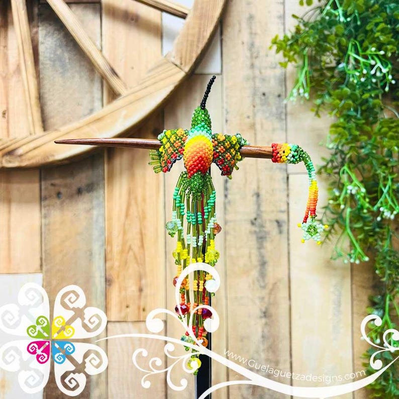 May include: A handcrafted beaded hummingbird sculpture with vibrant colors. The bird features a green body, a yellow and orange chest, and a long, beaded tail. It is mounted on a wooden dowel, with a wooden wheel and greenery in the background.