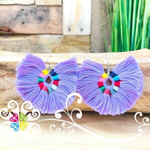 May include: A pair of lavender tassel earrings with a colorful circular design in the center. The earrings have a silver hook and are displayed on a wooden surface. The earrings are a bohemian style.