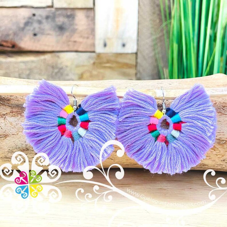 May include: A pair of lavender tassel earrings with a colorful circular design in the center. The earrings have a silver hook and are displayed on a wooden surface. The earrings are a bohemian style.