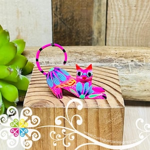 May include: A hand-painted wooden cat figurine with a pink body, blue and orange floral details, and black eyes. The cat is lying down with its tail curled up.
