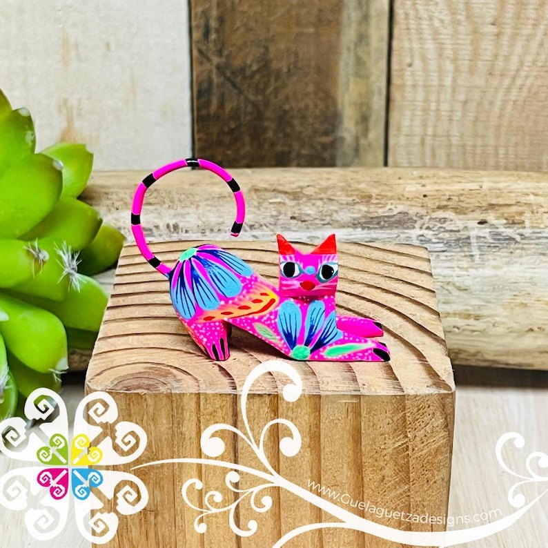 May include: A hand-painted wooden cat figurine with a pink body, blue and orange floral details, and black eyes. The cat is lying down with its tail curled up.