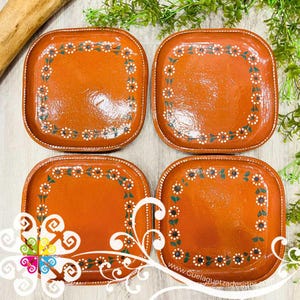 May include: Four square terracotta-colored plates with rounded corners. Each plate features a hand-painted floral border with white and yellow flowers and green leaves. The plates are arranged on a light-colored surface.