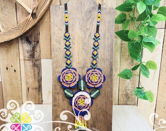 May include: A handcrafted beaded necklace featuring large blue and orange floral designs. The necklace has a black beaded chain with yellow and green accents. The necklace is displayed on a wooden stand against a rustic wood background.