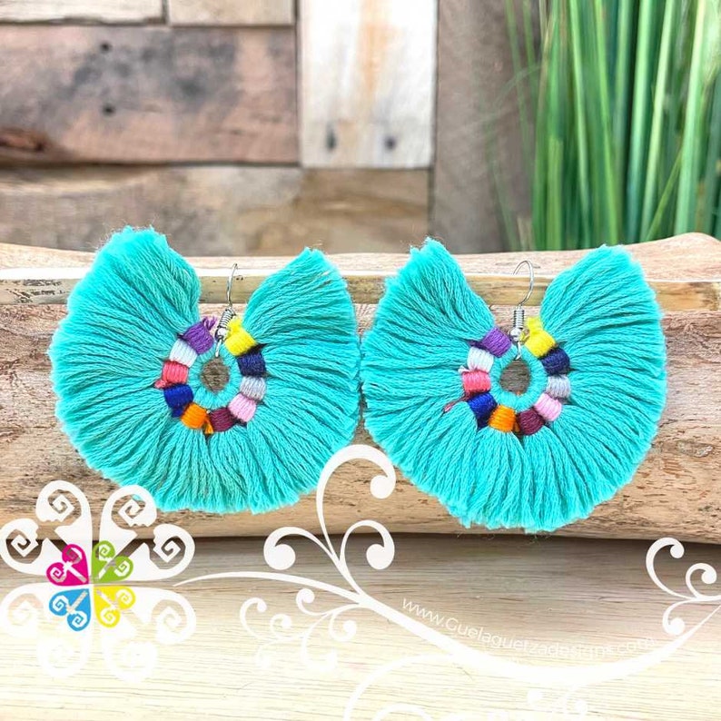 May include: A pair of turquoise tassel earrings. Each earring features a fan-shaped design with a central ring of colorful beads. The earrings are displayed on a wooden surface, showcasing their vibrant colors and intricate details.