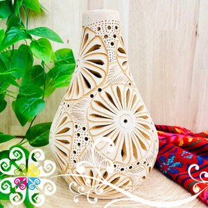 May include: Cream-colored ceramic vase with intricate floral cut-out designs. The vase has a bulbous body and a slender neck, with a series of small holes near the top. The design features a repeating pattern of stylized flowers. The vase is approximately 10 inches tall.