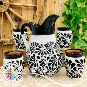 May include: A ceramic pitcher and four matching cups featuring a black and white floral pattern. The pitcher has a curved handle and a wide spout. Each piece is decorated with hand-painted black petals on a white background. The interior is terracotta.