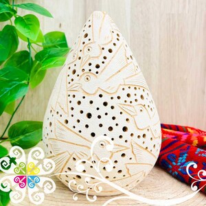 May include: Cream-colored, teardrop-shaped ceramic sculpture with carved bird designs and numerous small holes. The sculpture is displayed on a wooden surface, with green foliage and a colorful textile in the background.