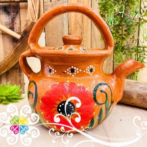 May include: Hand-painted terracotta teapot with a large red flower design. The teapot has a curved handle, a spout, and a lid. The design includes white and black accents, and green leaves. The teapot is a warm orange color.