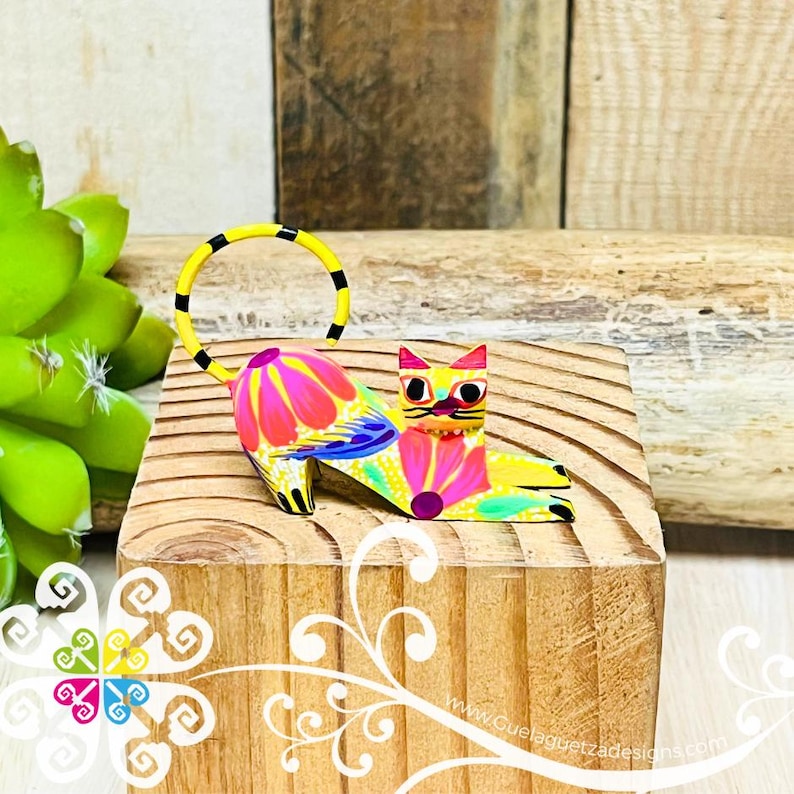 May include: A colorful wooden cat figurine with a yellow body, pink and blue flowers, and a black and yellow tail. The cat is lying down with its head turned to the side. The figurine is on a wooden surface.