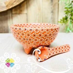 May include: Orange ceramic bowl and spoon set. The bowl has three small feet and is covered in white and blue polka dots. The spoon has a rounded bowl and a handle, also decorated with white and blue dots. The set is handmade.