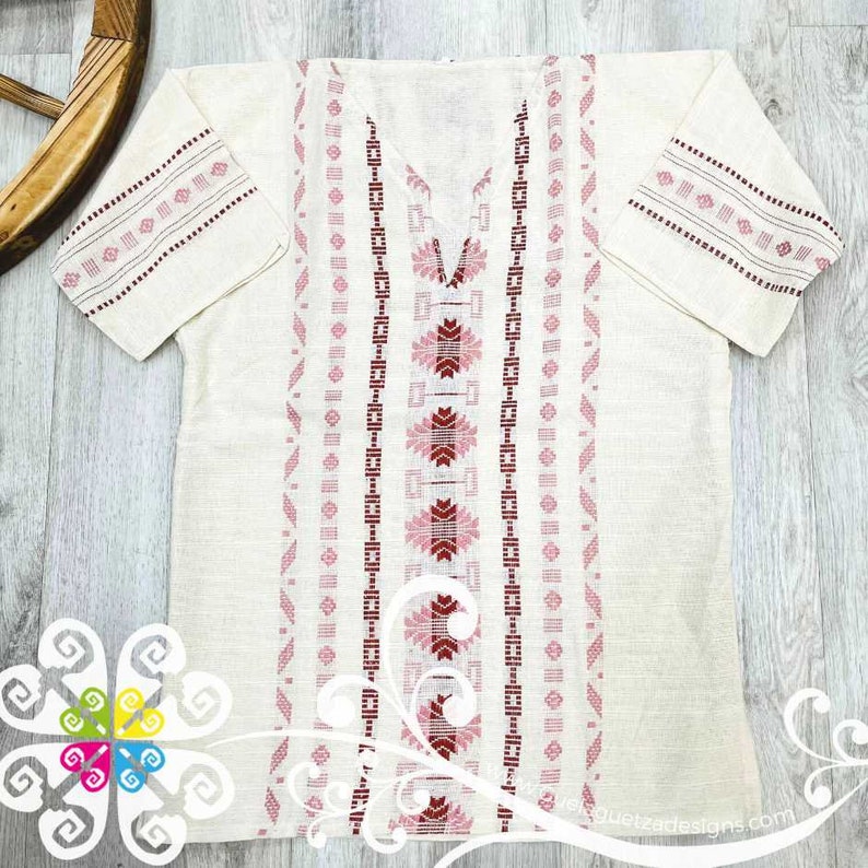 May include: A white linen shirt with pink and red embroidered geometric patterns on the sleeves and front. The shirt has a V-neck and short sleeves.