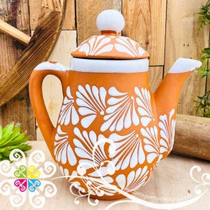 May include: Orange ceramic teapot with white floral design. The teapot has a white lid and handle. The spout and handle are also white. The teapot is sitting on a wooden surface.