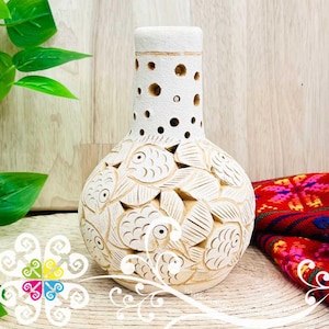 May include: A cream-colored, handcrafted ceramic vase with a carved fish and leaf design. The vase has a cylindrical neck with circular cutouts and a rounded body. The background includes a wooden surface and a colorful textile.
