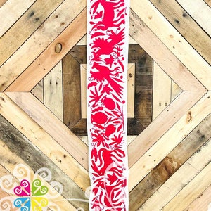 May include: A long, white textile strip with a vibrant red floral and animal design. The intricate pattern features stylized birds, leaves, and other organic shapes. The background is a wooden surface with a geometric pattern.
