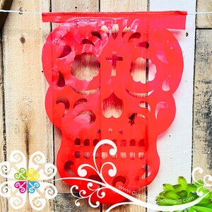 K&ouml;nnte beinhalten: Ein leuchtend roter Papierschnitt eines Zucker Sch&auml;dels mit filigranen Designs. Der Sch&auml;del hat ein Kreuz auf der Stirn und florale Muster um die Augen. Der Sch&auml;del h&auml;ngt an einem Holzzaun mit einer wei&szlig;en Schnur.