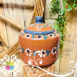 May include: A terracotta-colored ceramic jar with a lid, adorned with a floral pattern in white, black, and teal. The jar has a round shape and a gray-blue knob on the lid. The website www.Guelaguetzadesigns.com is visible.