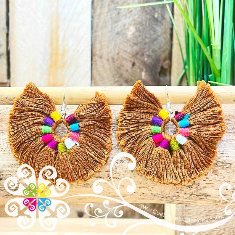 May include: A pair of brown tassel earrings with a fan-like shape. The earrings feature a colorful beaded center with pink, purple, blue, and green accents. The earrings are displayed on a wooden surface.
