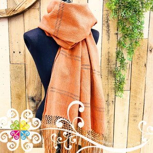 May include: A peach-colored scarf with a fringed edge and decorative cut-out details. The scarf is draped over a black mannequin, showcasing its woven texture and design. The background features a wooden plank wall and hanging greenery.