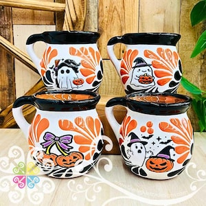 May include: Four white ceramic mugs with black rims and handles, featuring hand-painted orange and black floral patterns. Each mug displays unique Halloween-themed illustrations including ghosts, jack-o'-lanterns, and bows. The text www.quelaguetzadesigns.com is visible.