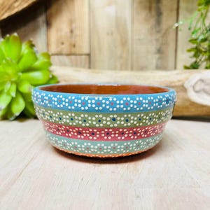 May include: Hand-painted ceramic bowl with a colorful design. The bowl features bands of blue, green, red, and orange, each adorned with white and blue dots. The interior is a warm brown color, and the bowl is set against a wooden background.