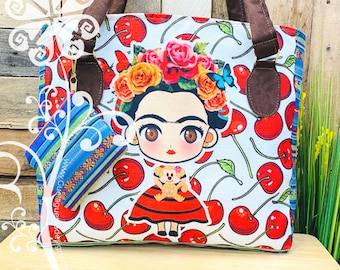 May include: Tote bag featuring a Frida Kahlo illustration with a floral crown, surrounded by red cherries on a white background. Includes a matching small pouch with a colorful striped pattern. The bag has brown handles.