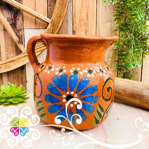 May include: Hand-painted terracotta pitcher with a large blue floral design. The pitcher has a handle and is decorated with white dots and green leaves. The background is a wooden surface with greenery.