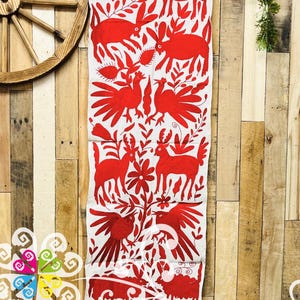 May include: A white textile runner with a repeating red pattern of stylized animals, birds, and floral elements. The design is symmetrical and covers the entire length of the runner. The runner is displayed against a wooden background.