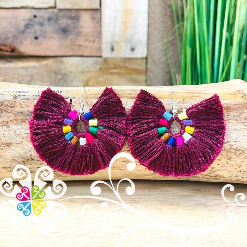 May include: A pair of burgundy tassel earrings with a fan-like shape. The earrings feature a circular design with colorful accents and silver-tone ear wires. The earrings are displayed on a wooden surface.