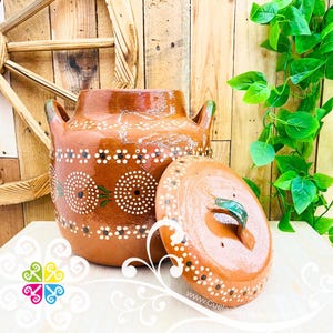 May include: A brown ceramic pot with a lid, featuring white and black dot and floral patterns. The pot has two handles and a green accent on the lid handle. The background includes wooden planks and green foliage.