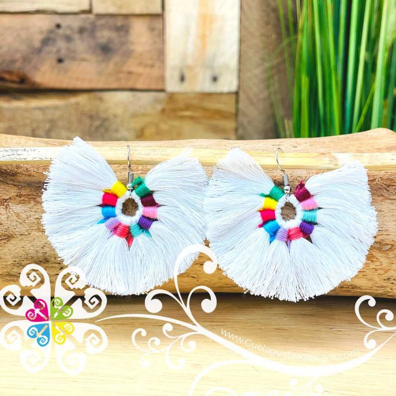 May include: A pair of white tassel earrings with a colorful center design. The earrings have a fan-like shape with a rainbow of colors in the center, including red, yellow, green, and purple. The earrings are displayed on a wooden surface.